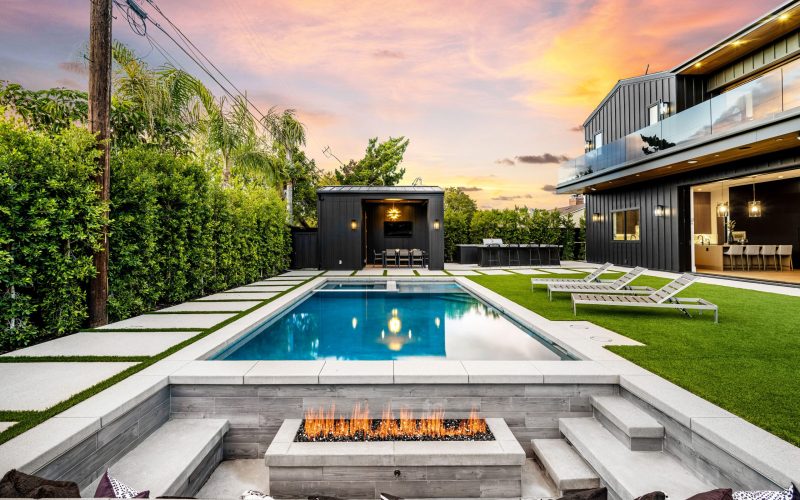 Modern backyard with a swimming pool, fire pit, and lounge chairs at sunset, featuring a contemporary house with large windows and manicured garden.