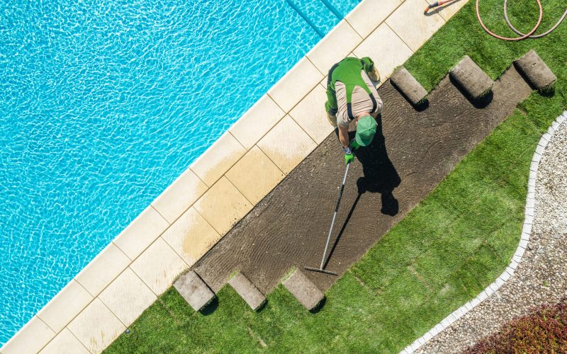 Caucasian Professional Landscaper Installing Brand New Grass Turfs Around Residential Swimming Pool. Raking Soil Before Installation. Gardening and Landscaping Industry Theme.